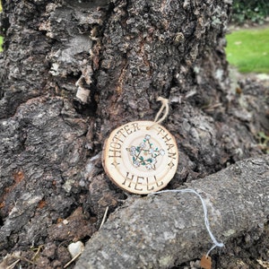 Puede incluir: Un adorno redondo de madera con el texto "Hotter Than Hell" y un pentagrama con flores blancas en su interior. El adorno está colgado de un tronco de árbol.