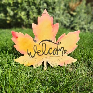 May include: A wooden welcome sign shaped like a maple leaf with a watercolor-like orange and yellow design. The word "welcome" is written in black calligraphy.