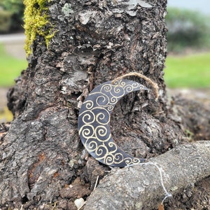 Puede incluir: Un adorno de media luna negro con un diseño de espiral dorado. El adorno está colgado de un tronco de árbol.