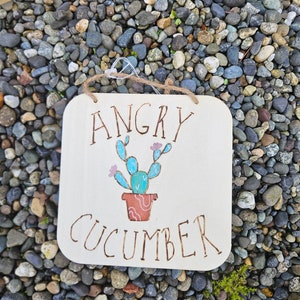 Puede incluir: Un letrero de madera blanco con el texto "Angry Cucumber" escrito en negro. Un cactus azul con flores rosas está dibujado en el centro del letrero.