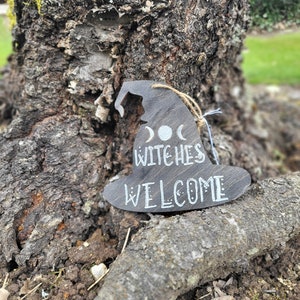 Puede incluir: Un adorno de sombrero de bruja de madera con el texto "Witches Welcome" y tres lunas pintadas.