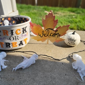 May include: A white bucket with the words "Trick or Treat" in black and orange letters. A white pumpkin with the words "Trick or Treat" in black letters. A fall leaf with the word "Welcome" in black letters. A string of white ghosts.