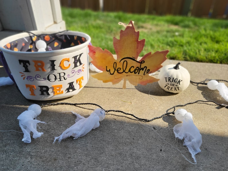 May include: A white bucket with the words "Trick or Treat" in black and orange letters. A white pumpkin with the words "Trick or Treat" in black letters. A fall leaf with the word "Welcome" in black letters. A string of white ghosts.