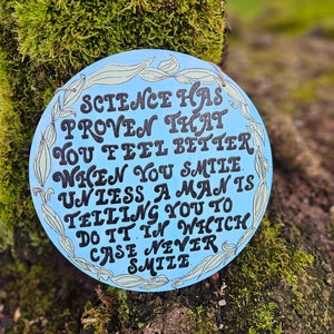 May include: A blue round sign with white floral accents and black text that reads "Science has proven that you feel better when you smile unless a man is telling you to do it in which case never smile."
