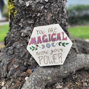 Puede incluir: Un letrero de madera hexagonal con el texto "You are magical know your power" en letras rosa, azul y verde. El letrero está decorado con un pequeño diseño floral y tres lunas.