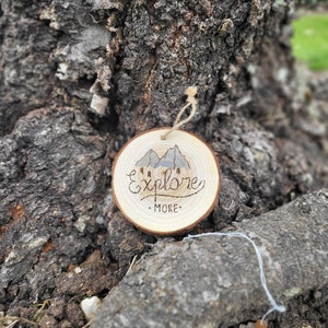 Puede incluir: Adorno redondo de madera con las palabras "Explore More" grabadas. El adorno presenta un diseño de cordillera y un lazo de cuerda para colgar. El adorno está colocado contra un tronco de árbol.