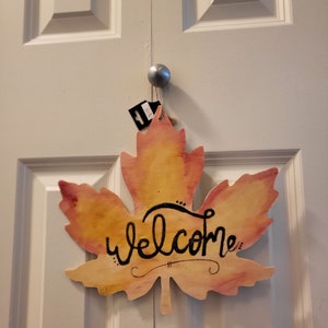 May include: A wooden maple leaf-shaped welcome sign with a watercolor-like design in shades of yellow, orange, and pink. The word "welcome" is written in black cursive lettering.