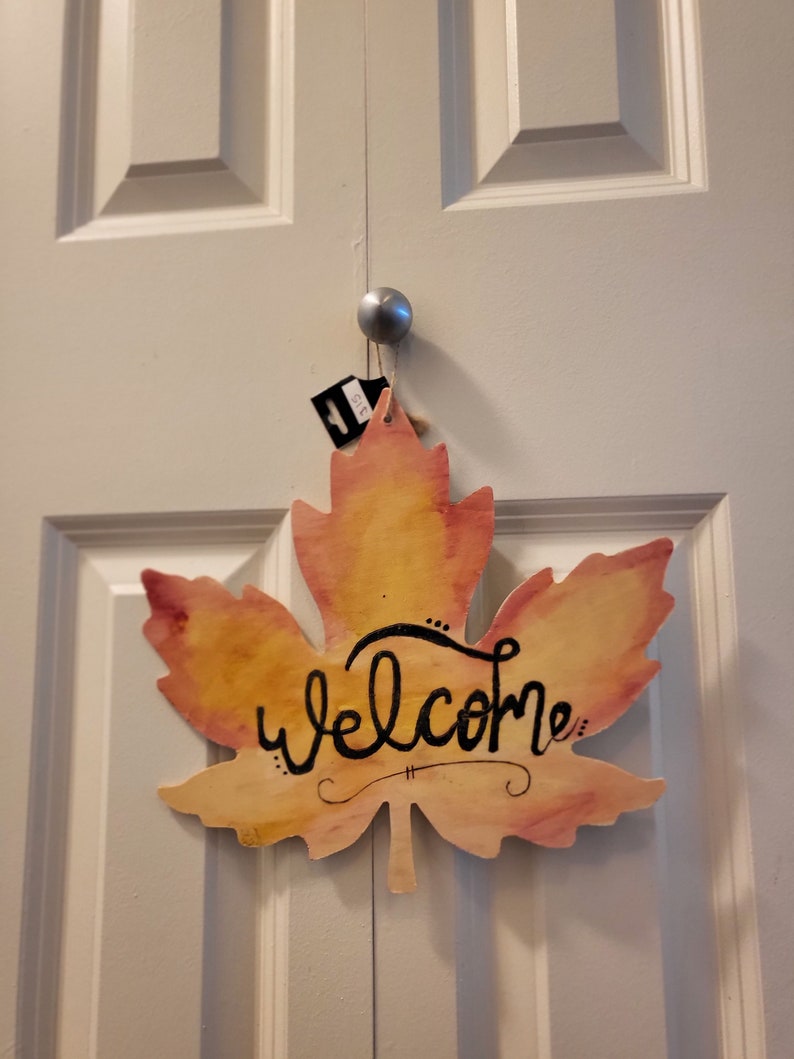 May include: A wooden maple leaf-shaped welcome sign with a watercolor-like design in shades of yellow, orange, and pink. The word "welcome" is written in black cursive lettering.