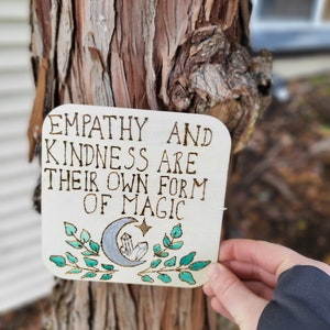 Puede incluir: Un letrero de madera con el texto "Empatía y amabilidad son su propia forma de magia" en letras marrones. El letrero está decorado con una luna creciente, estrellas y hojas verdes.
