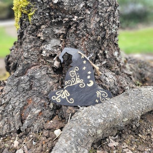 Puede incluir: Un adorno de sombrero de bruja negro con detalles pintados en oro, incluyendo estrellas, lunas y espirales. El adorno está colgado en un tronco de árbol.