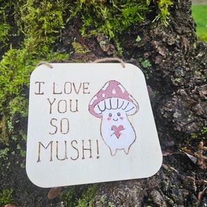 Puede incluir: Un letrero de madera con el texto "I love you so mush!" y un hongo de dibujos animados sosteniendo un corazón. El hongo es rosa y blanco con un corazón rojo.
