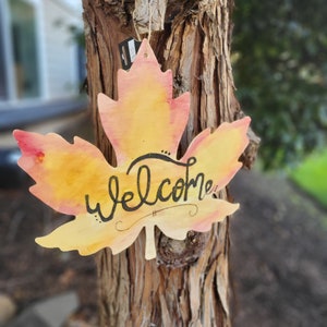 May include: A wooden maple leaf painted in shades of yellow and orange with the word "welcome" written in black calligraphy. The leaf is hanging on a tree trunk.