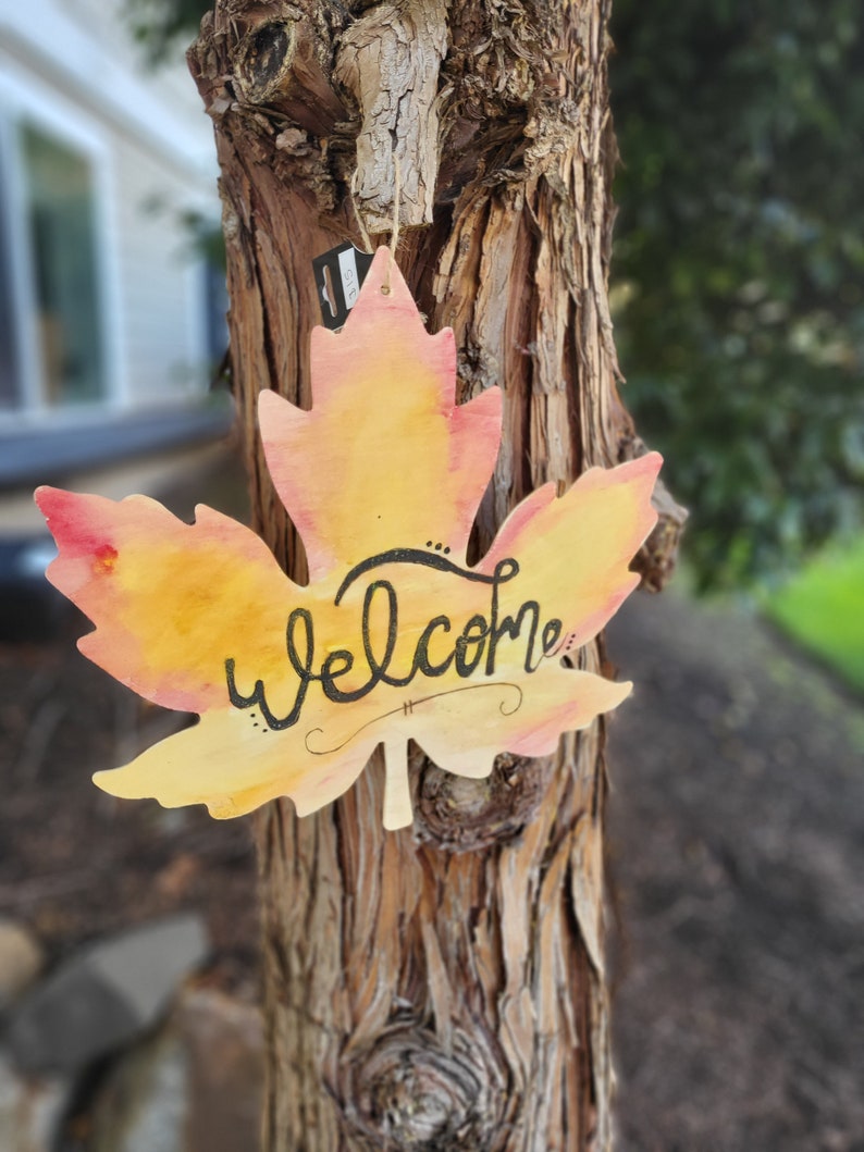 May include: A wooden maple leaf painted in shades of yellow and orange with the word "welcome" written in black calligraphy. The leaf is hanging on a tree trunk.