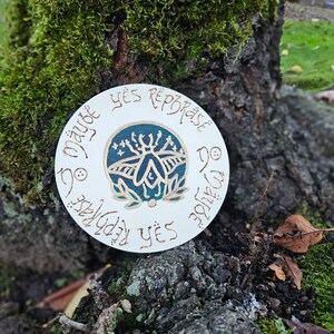 May include: A round, off-white decorative piece with the words "Yes Reprobate" and "Maybe No" in a circular pattern. The center features a teal and gold insect design. The piece is set against a mossy tree trunk.