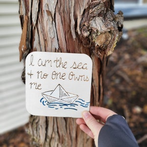 Puede incluir: Un letrero de madera blanco con letras doradas que dice "Soy el mar y nadie me posee". Debajo del texto hay un dibujo simple de un barco de papel en olas azules.