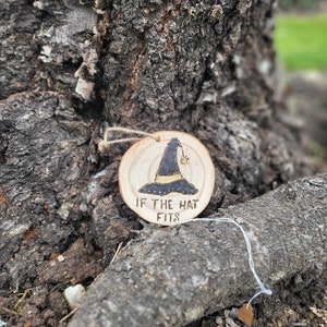 Puede incluir: Un adorno redondo de madera con un diseño de sombrero de bruja y el texto "Si el sombrero encaja".