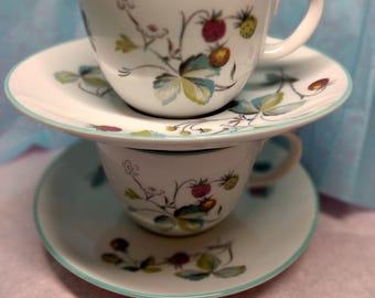 Puede incluir: Un juego de dos tazas de té y platillos blancos con un patrón floral con hojas verdes y fresas rojas. Los platillos tienen un borde azul claro.
