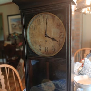 May include: A rectangular dark wood wall clock featuring a circular face with a weathered, off-white background and black Arabic numerals. The clock hands are black, and the lower section contains a glass panel revealing a pendulum inside.