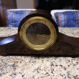 May include: A dark brown wooden mantel clock with a horizontal, elongated base and a central circular face. The clock features a gold-toned bezel, a cream-colored dial with black Arabic numerals, and two gold-colored hands behind a glass cover.