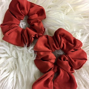 May include: Two rust-colored scrunchies, one with a bow and one without. The scrunchies are made of soft fabric and are perfect for holding hair in place.