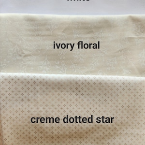 May include: Three fabric swatches: white, ivory floral, and creme dotted star. The white fabric is plain. The ivory fabric has a floral pattern. The creme fabric has a dotted star pattern. The names of the fabrics are written on the fabric.