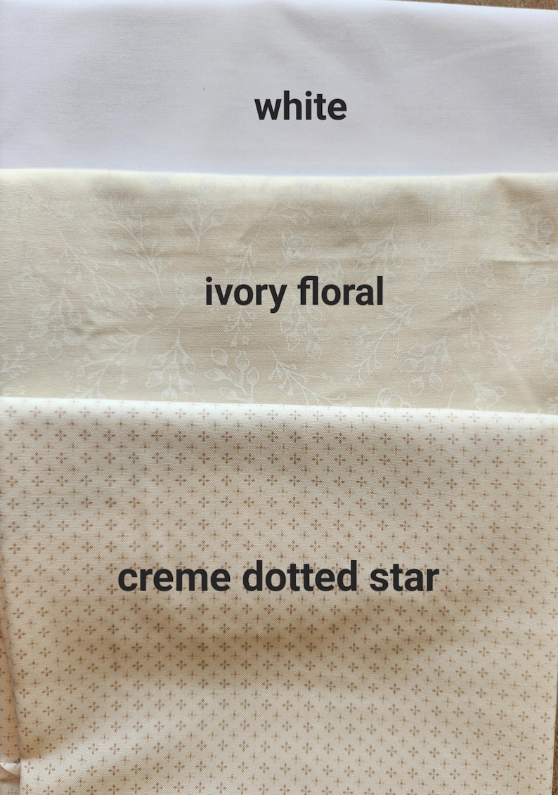 May include: Three fabric swatches: white, ivory floral, and creme dotted star. The white fabric is plain. The ivory fabric has a floral pattern. The creme fabric has a dotted star pattern. The names of the fabrics are written on the fabric.