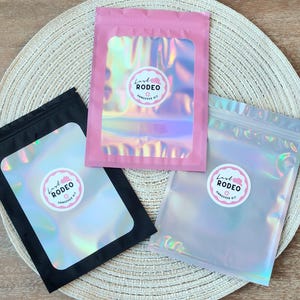 May include: Three iridescent pouches in pink, black, and clear, each with a circular sticker that reads "Last Rodeo Hangover Kit." The pouches are arranged on a woven placemat, with the pink pouch centered.