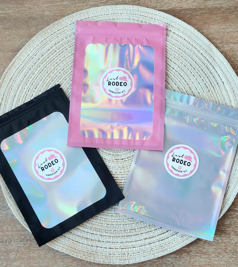 May include: Three iridescent pouches in pink, black, and clear, each with a circular sticker that reads "Last Rodeo Hangover Kit." The pouches are arranged on a woven placemat, with the pink pouch centered.