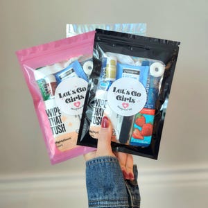 May include: Two clear plastic zip bags, one pink and one black, filled with small items. The pink bag has the text "Let's Go Girls" and "Wipe That Tush". The black bag also has the text "Let's Go Girls". The bags are held by a person.