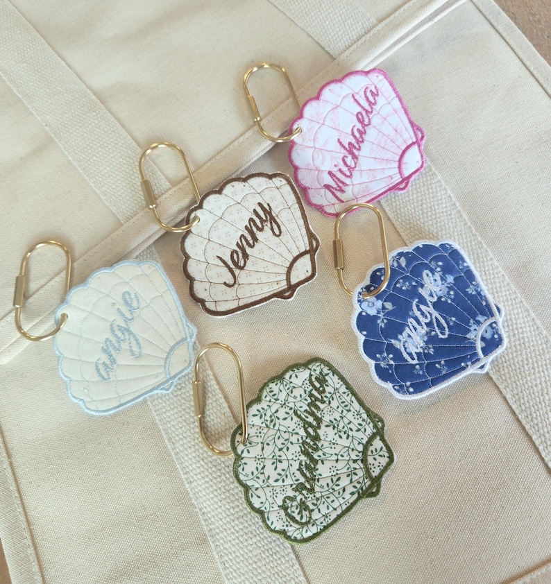 May include: Five seashell-shaped keychains with gold-colored clasps. Each keychain features a different design and name: "Angie", "Jenny", "Michaela", "Grandma", and a blue floral pattern. The keychains are resting on a beige tote bag.