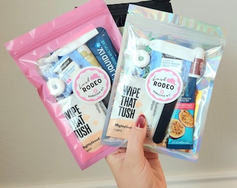May include: Two clear plastic bags filled with various small items, including wipes, lip balm, and electrolyte mix. One bag is pink, the other is iridescent. Each bag has a sticker that says "Last Rodeo Hangover Kit" and "Wipe That Tush."