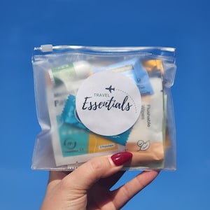 May include: A clear, zippered pouch filled with travel-sized essentials. A white circular label with the text "Travel Essentials" and a small airplane graphic is affixed to the pouch. The background is a clear blue sky.