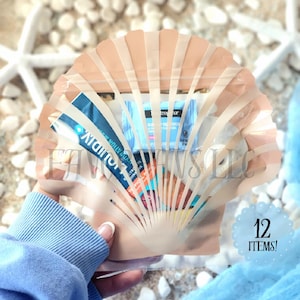 May include: A clear plastic seashell-shaped pouch with a zipper closure. The pouch is filled with various small items, including a blue and white tube of sunscreen, a blue and white bar of soap, and a small white bottle. The pouch is held in a hand against a background of white sand and seashells. The text "12 ITEMS!" is printed on a blue circle in the lower right corner of the image.