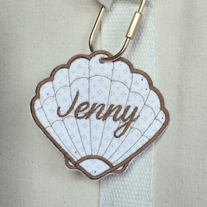 May include: A seashell-shaped tag with the name "Jenny" embroidered in brown script. The tag has a white background with a subtle floral pattern and a brown border. It is attached to a beige tote bag with a gold carabiner clip.
