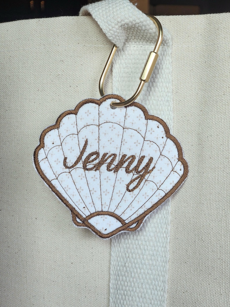 May include: A seashell-shaped tag with the name "Jenny" embroidered in brown script. The tag has a white background with a subtle floral pattern and a brown border. It is attached to a beige tote bag with a gold carabiner clip.