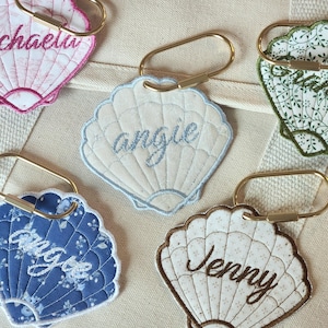 May include: Embroidered seashell-shaped keychains in various colors, each with a name stitched in cursive. The keychains have gold-colored metal clasps. The names include Angie, Jenny, and Michaela. The keychains are on a beige background.