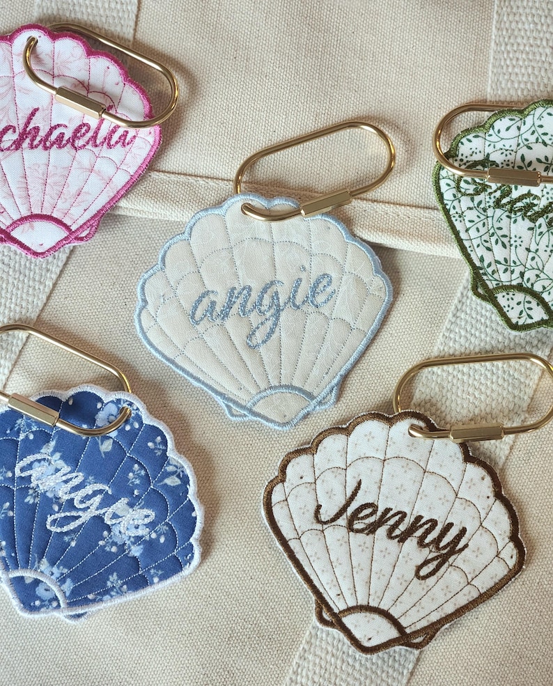 May include: A collection of seashell-shaped keychains with embroidered names. The keychains feature various colors and patterns, including pink, blue, and floral designs. Each keychain has a gold-colored metal clasp.