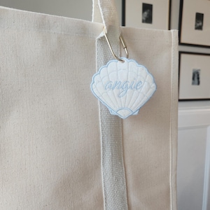 May include: A cream-colored tote bag with a light blue seashell-shaped tag that reads "angie" in cursive. The tag is attached to the bag's handle with a metal clasp. The bag has a light gray stripe down the front.