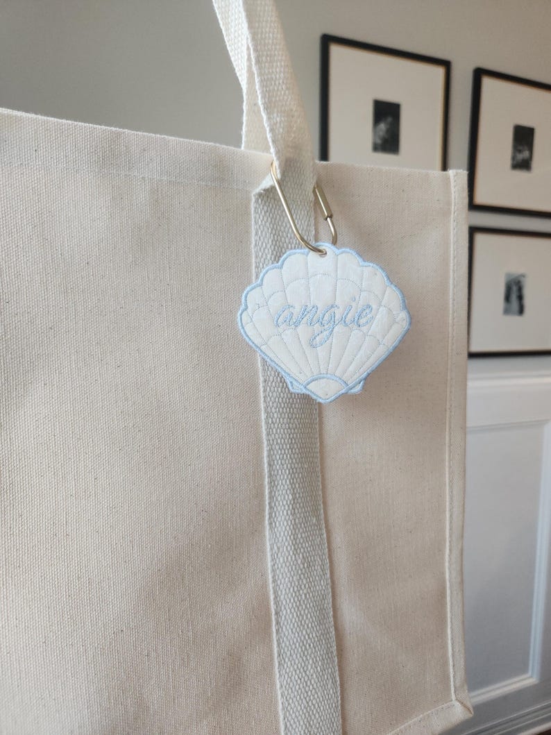 May include: A cream-colored tote bag with a light blue seashell-shaped tag that reads "angie" in cursive. The tag is attached to the bag's handle with a metal clasp. The bag has a light gray stripe down the front.