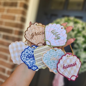 May include: A collection of seashell-shaped keychains in various patterns and colors, including gingham, floral, and striped designs. Each keychain features a name in cursive script and a gold-colored ring for attachment.