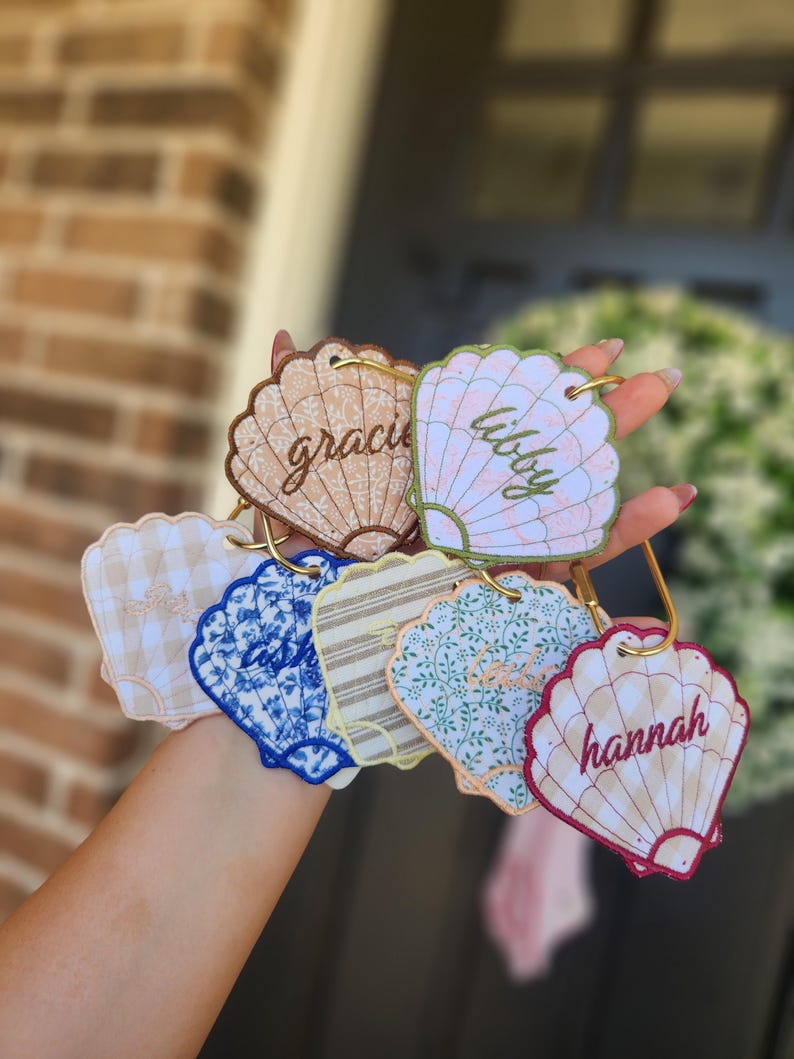 May include: A collection of seashell-shaped keychains in various patterns and colors, including gingham, floral, and striped designs. Each keychain features a name in cursive script and a gold-colored ring for attachment.