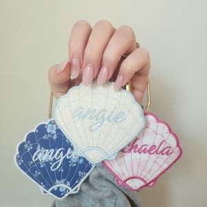 May include: Three seashell-shaped embroidered name tags. One is blue with a floral pattern and the name "Angie". Another is white with the name "Angie". The third is pink with the name "Michaela".