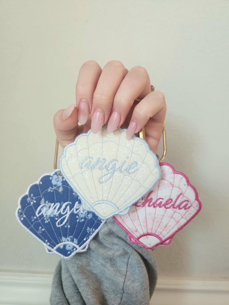 May include: Three seashell-shaped embroidered name tags. One is blue with a floral pattern and the name "Angie". Another is white with the name "Angie". The third is pink with the name "Michaela".