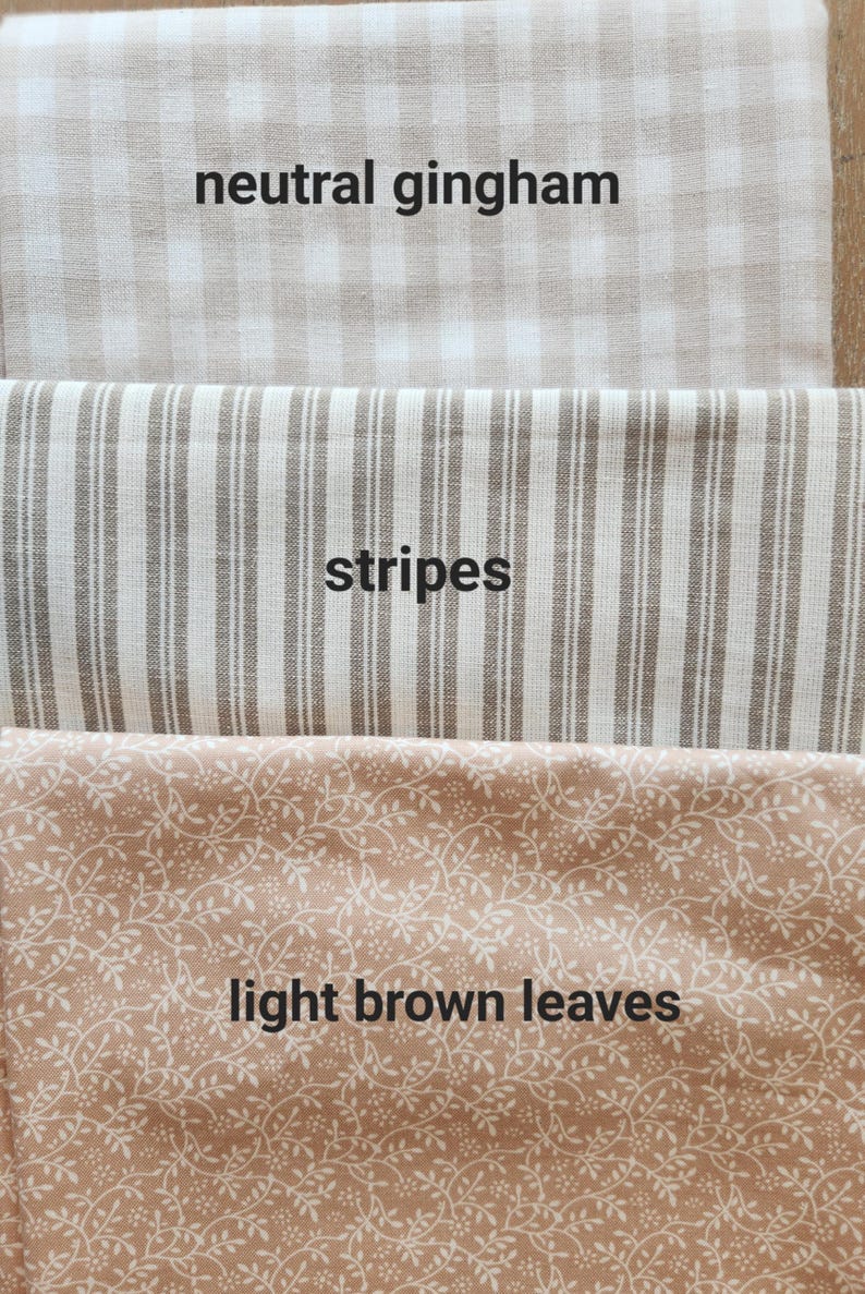 May include: Close-up of three fabric swatches. The top swatch is neutral gingham, the middle swatch has stripes, and the bottom swatch features light brown leaves. The text "neutral gingham", "stripes", and "light brown leaves" are printed on the fabric.