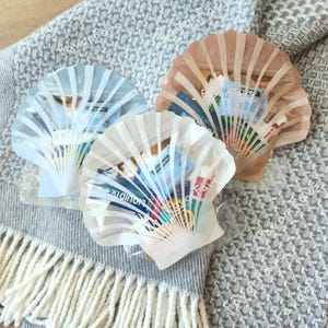 May include: Three seashell-shaped bags, each filled with various items, rest on a textured blue and gray blanket. The bags are in shades of white, light blue, and tan. The text "Favor Fous" is visible on the bags.