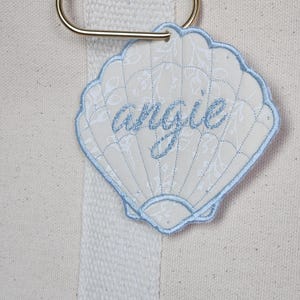 May include: A seashell-shaped tag with the name "angie" embroidered in light blue thread. The tag has a light blue border and is attached to a gold-colored metal ring. The background is a cream-colored canvas.