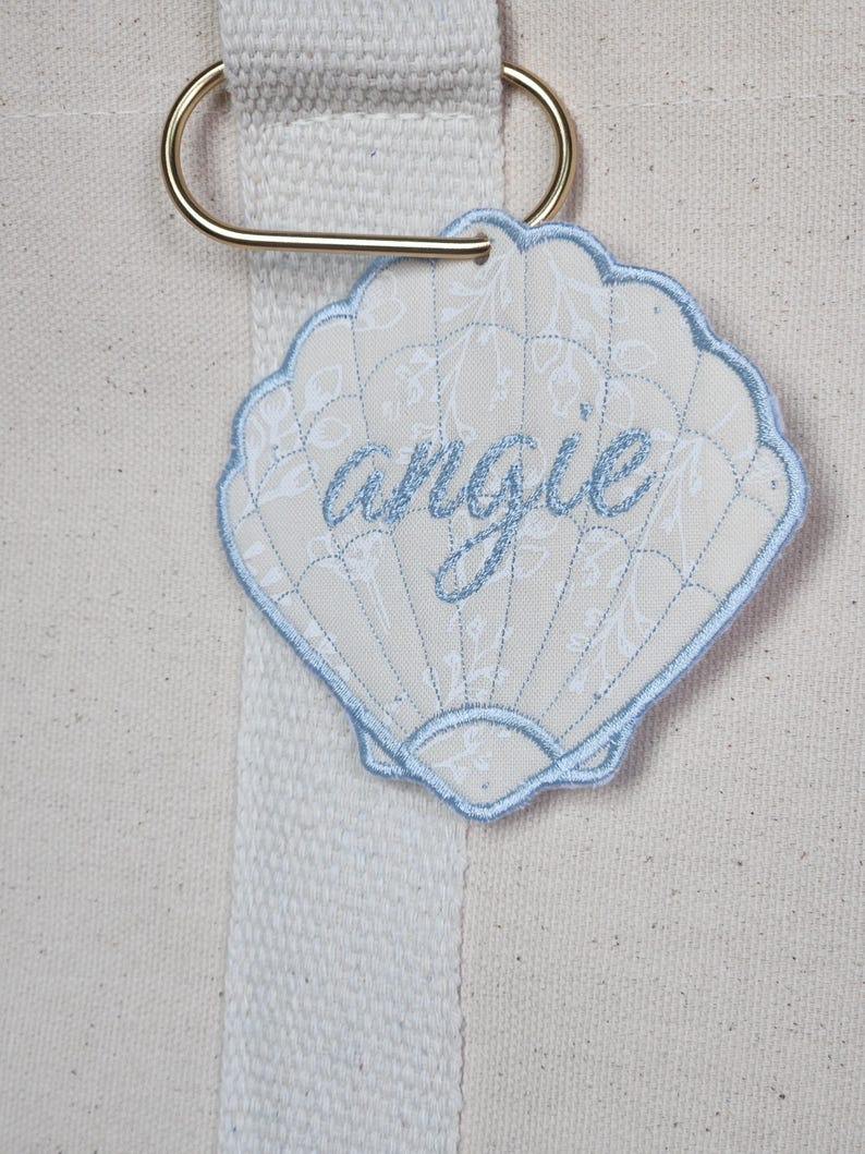 May include: A seashell-shaped tag with the name "angie" embroidered in light blue thread. The tag has a light blue border and is attached to a gold-colored metal ring. The background is a cream-colored canvas.