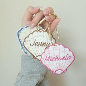 May include: Embroidered seashell keychains in white, brown, and pink, each with a name: "Jenny" and "Michaela". The keychains are attached to a gold-colored clasp. The background is a solid, light beige color.