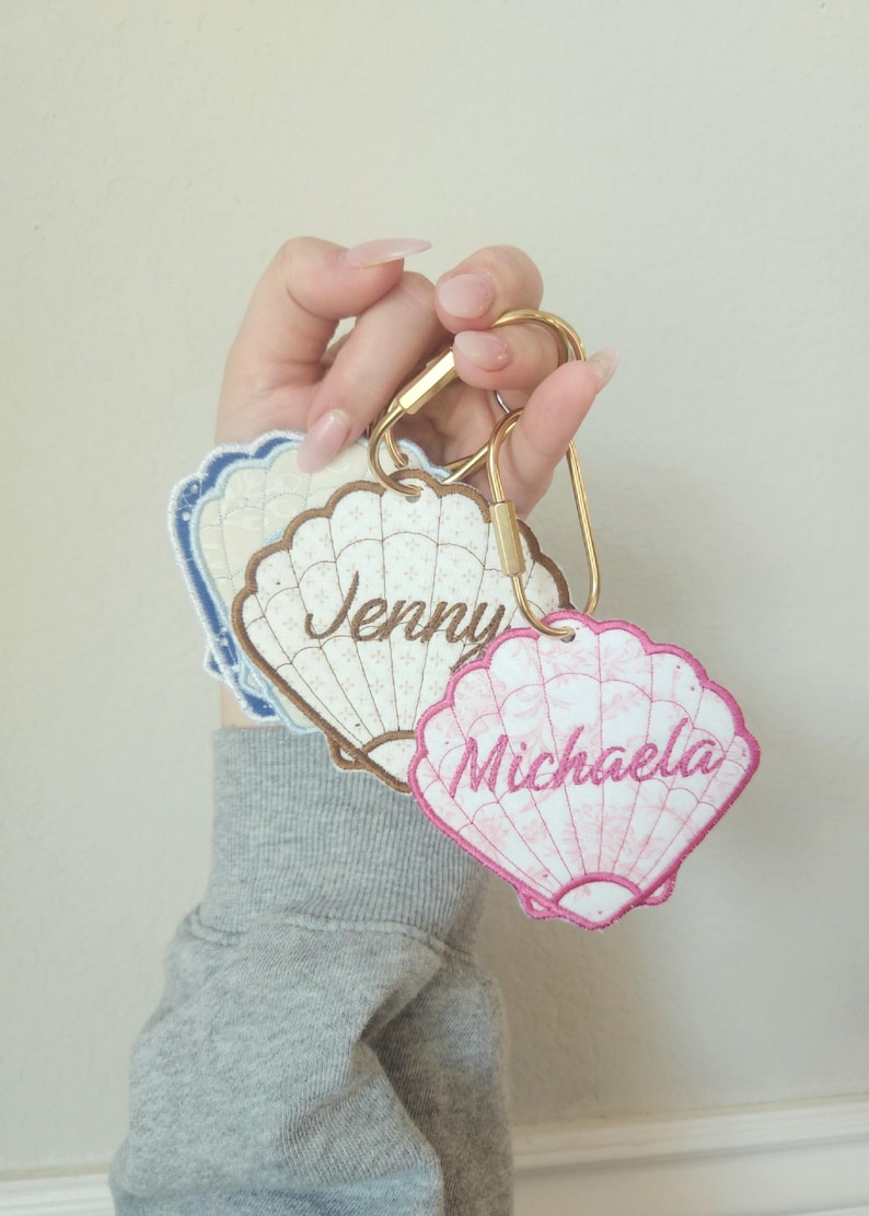 May include: Embroidered seashell keychains in white, brown, and pink, each with a name: "Jenny" and "Michaela". The keychains are attached to a gold-colored clasp. The background is a solid, light beige color.