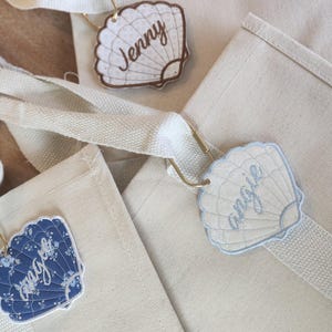 May include: Close-up of three canvas tote bags with seashell-shaped name tags. The tags are in various colors, including blue and cream, and feature names like "Jenny" and "Angie" in cursive script. The bags have cream-colored straps.